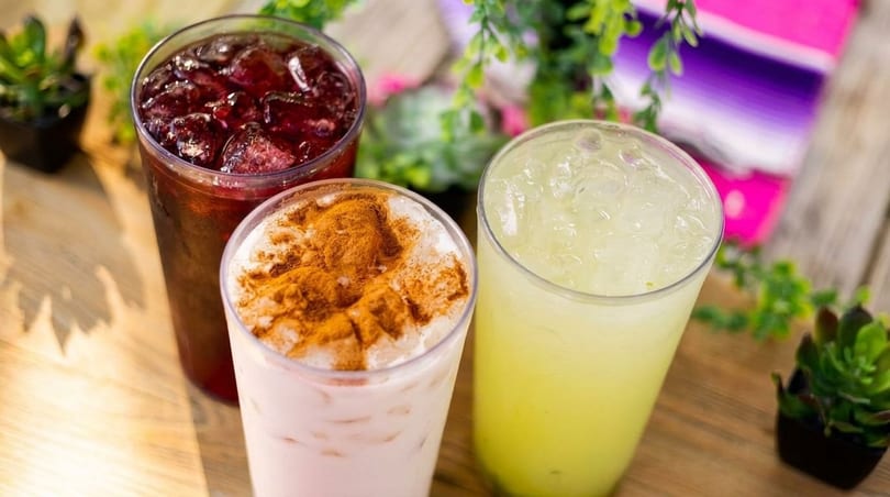 Three refreshing drinks: red, horchata with cinnamon, and light green, on a wooden surface.