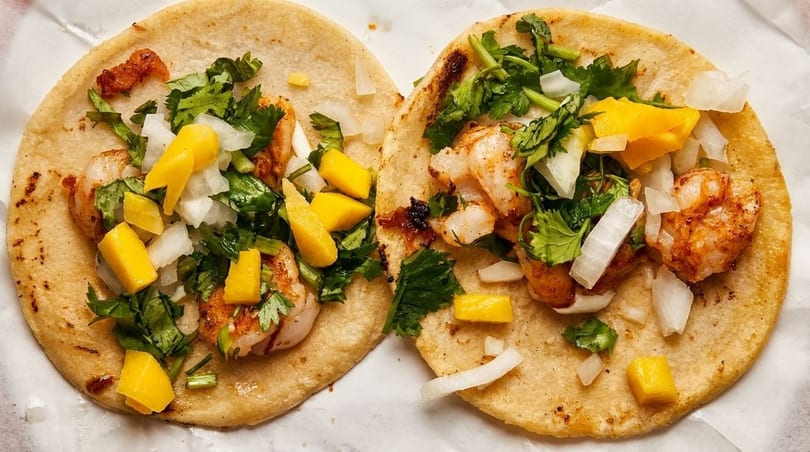 Two shrimp tacos topped with diced mango, onions, and cilantro on corn tortillas.