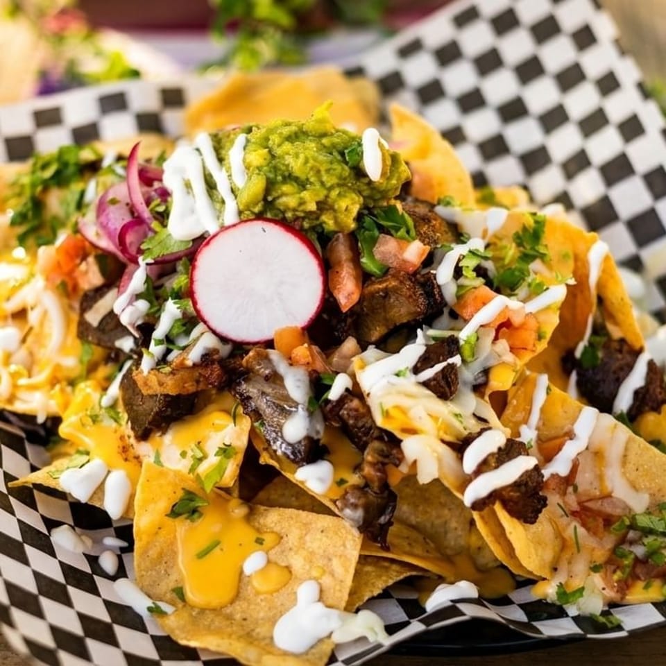 Loaded nachos with meat, cheese, guacamole, radish, and sour cream on a checkered paper.