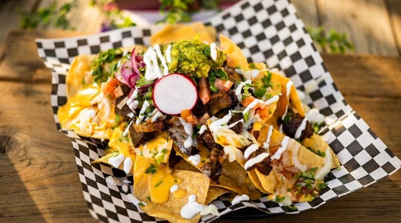 Loaded nachos with meat, cheese, guacamole, radish, and sour cream on a checkered paper.